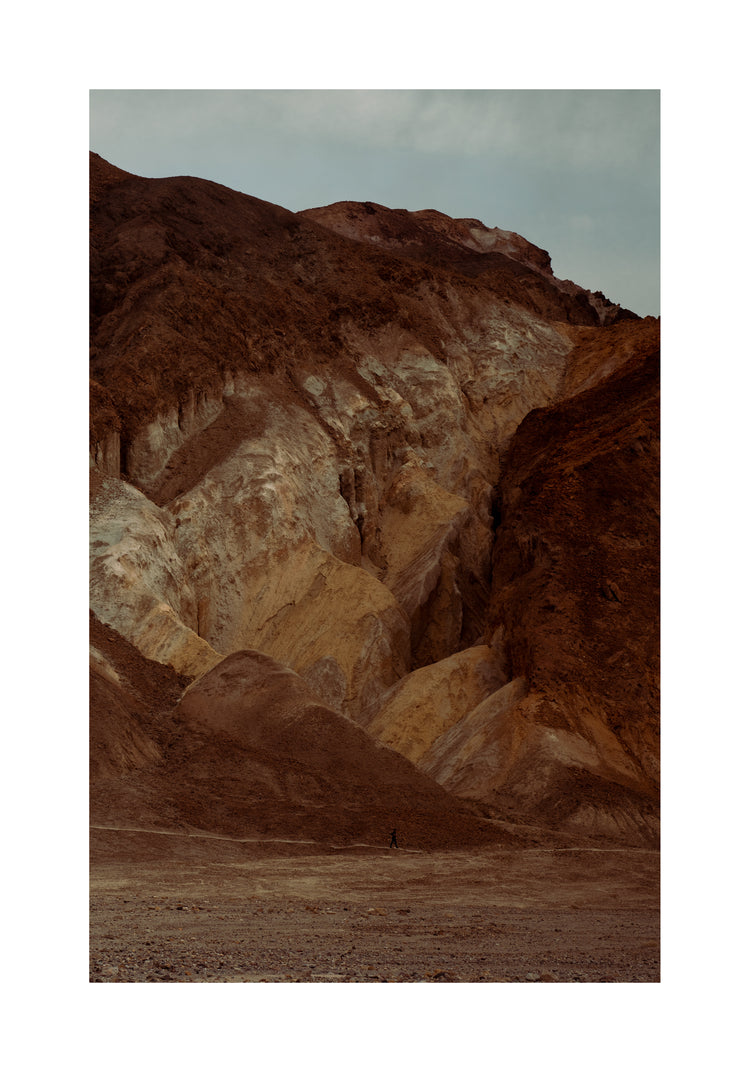 Tiny man walking along the bottom of a massive brown and beige mountain.
