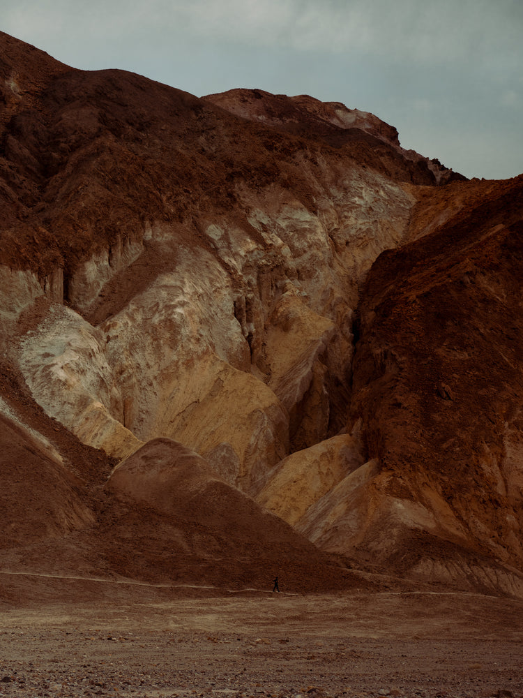 Tiny man walking along the bottom of a massive brown and beige mountain.