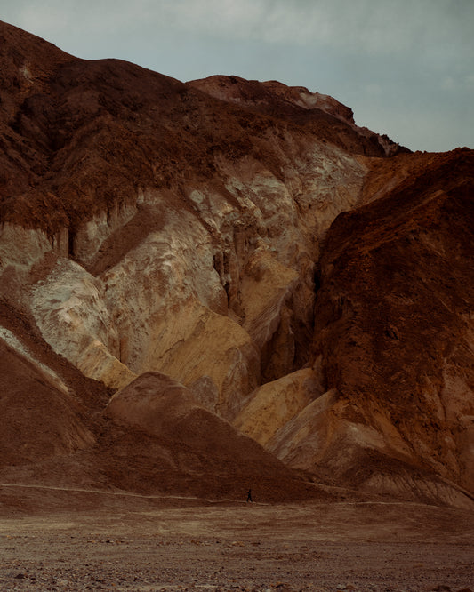 Tiny man walking along the bottom of a massive brown and beige mountain.
