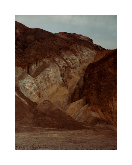 Tiny man walking along the bottom of a massive brown and beige mountain.