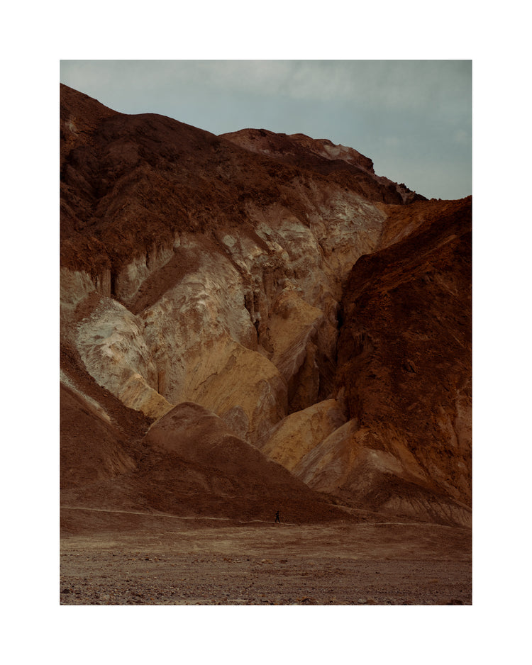Tiny man walking along the bottom of a massive brown and beige mountain.