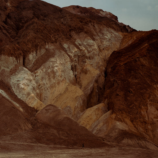 Tiny man walking along the bottom of a massive brown and beige mountain.