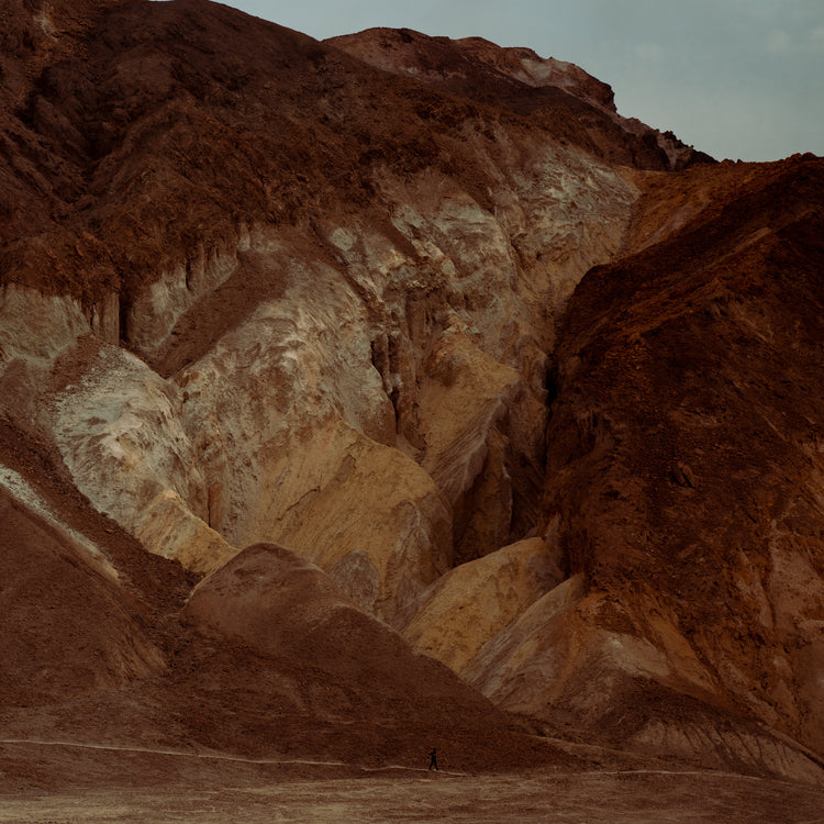 Tiny man walking along the bottom of a massive brown and beige mountain.