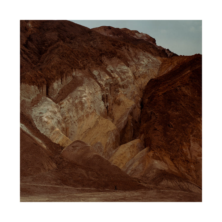 Tiny man walking along the bottom of a massive brown and beige mountain.