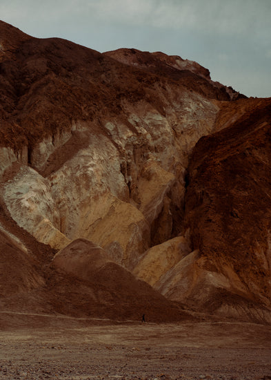 Tiny man walking along the bottom of a massive brown and beige mountain.