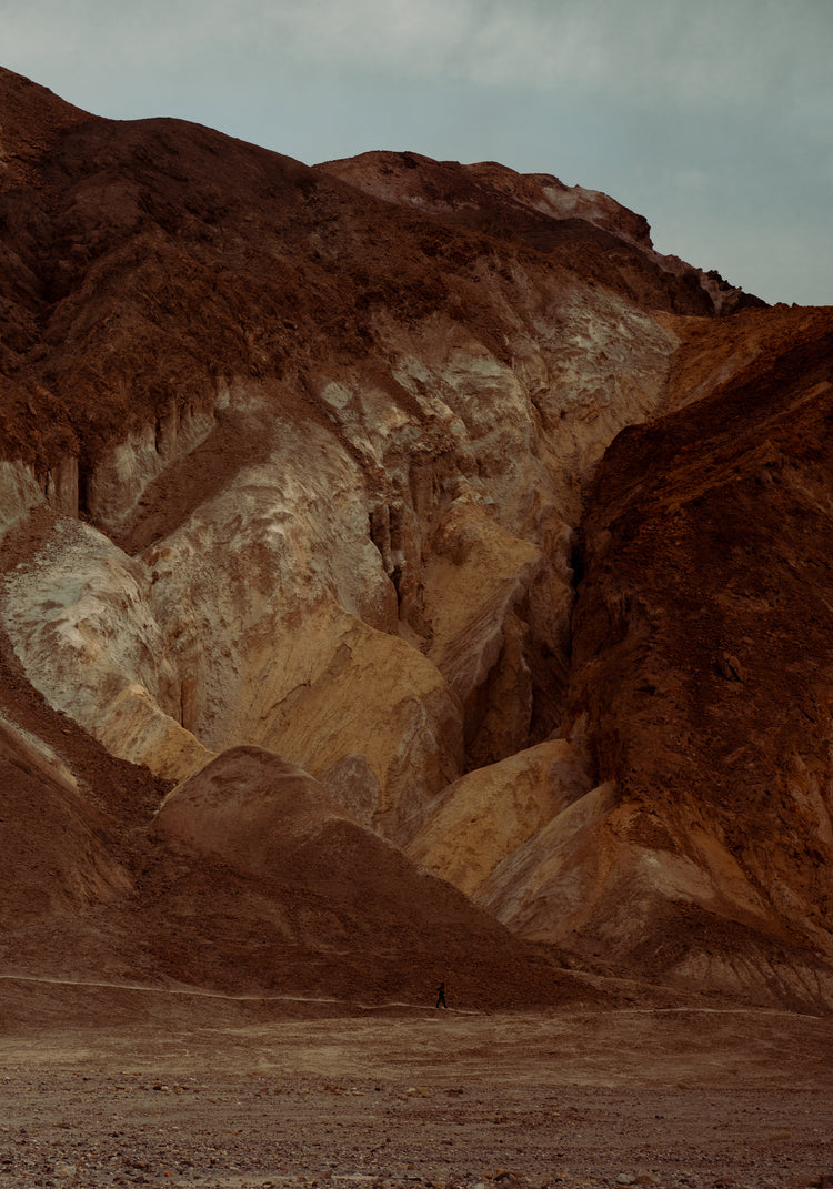 Tiny man walking along the bottom of a massive brown and beige mountain.