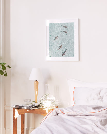A bright bedroom showcases a neatly made bed with a white pillow and comforter. A small wooden table beside it bears a lamp, water glass, and book. Above the bed, the On Patrol poster adds calmness, while subtle background music suggests an adventurous undertone.
