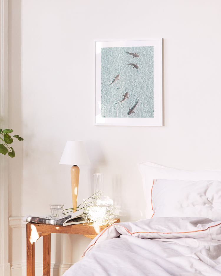 A bright bedroom showcases a neatly made bed with a white pillow and comforter. A small wooden table beside it bears a lamp, water glass, and book. Above the bed, the On Patrol poster adds calmness, while subtle background music suggests an adventurous undertone.