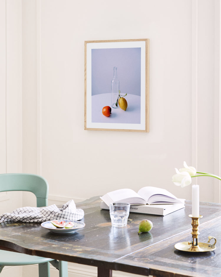 A dining table holds an open book, a glass of water, a candle, and a small plate with food next to a cloth napkin. A green chair is nearby. The Groupie Poster, depicting a bottle, lemon, and tomato, hangs on the wall. A white tulip in a vase decorates the table, swaying in the breeze from an unseen fan.