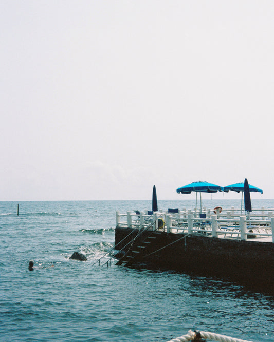 The Santa Marinella Poster showcases Italys sunlit coast with a concrete pier, blue umbrellas, and lounge chairs. A swimmer enjoys the serene blue water under a mostly clear sky dotted with clouds, capturing the essence of the enchanting Mediterranean seascapes.