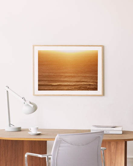 The Ikaros Poster, depicting a serene ocean sunset, adorns the wall above a minimalist wooden desk. The desk features a sleek white lamp, cup and saucer, and stacked books, while a light gray chair invites contemplation.