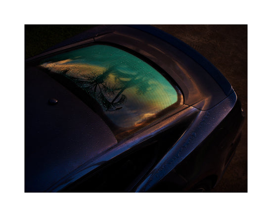 Sunset sky and outlines of palm trees reflected in a rain-stained car window.