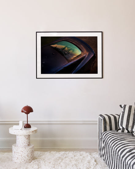 A modern living room boasts a striped sofa, a round white speckled side table with a brown lamp, and the Gas Up Poster, capturing the pedal to the metal spirit with car reflections of trees and sky on the cream-colored wall.