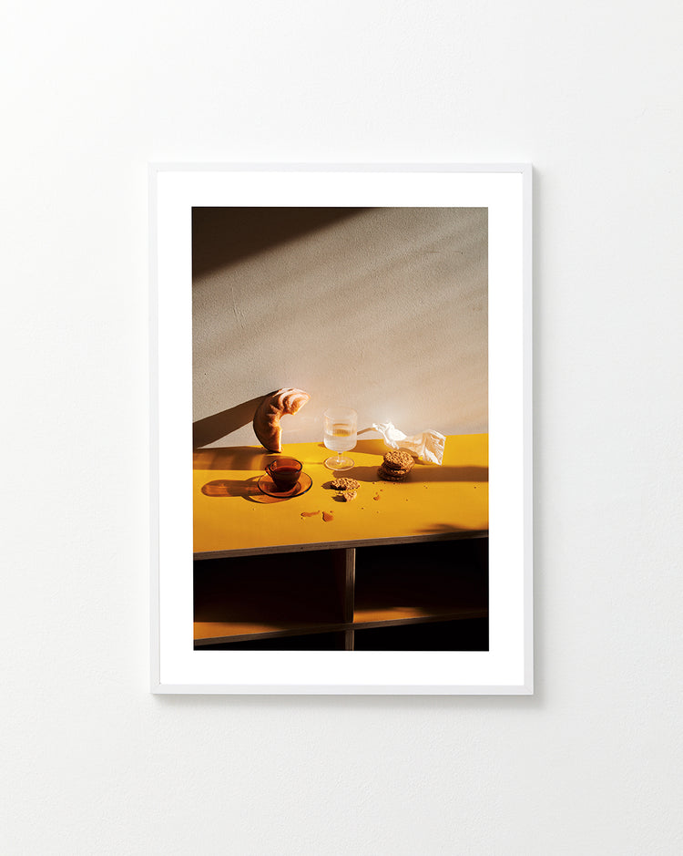 The Would You Like Some Coffee? poster depicts a still life on a white wall featuring a croissant, glass of water, wooden bowl, and cookies on a yellow shelf bathed in sunlight, with long shadows cast across the scene.