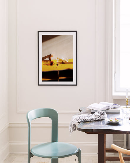 A minimalist room features a Would You Like Some Coffee? poster on a white wall, showing a hand pouring liquid into a cup on a yellow table. Nearby are a blue chair and dark wooden table with an open book and patterned cloth, all bathed in natural light.