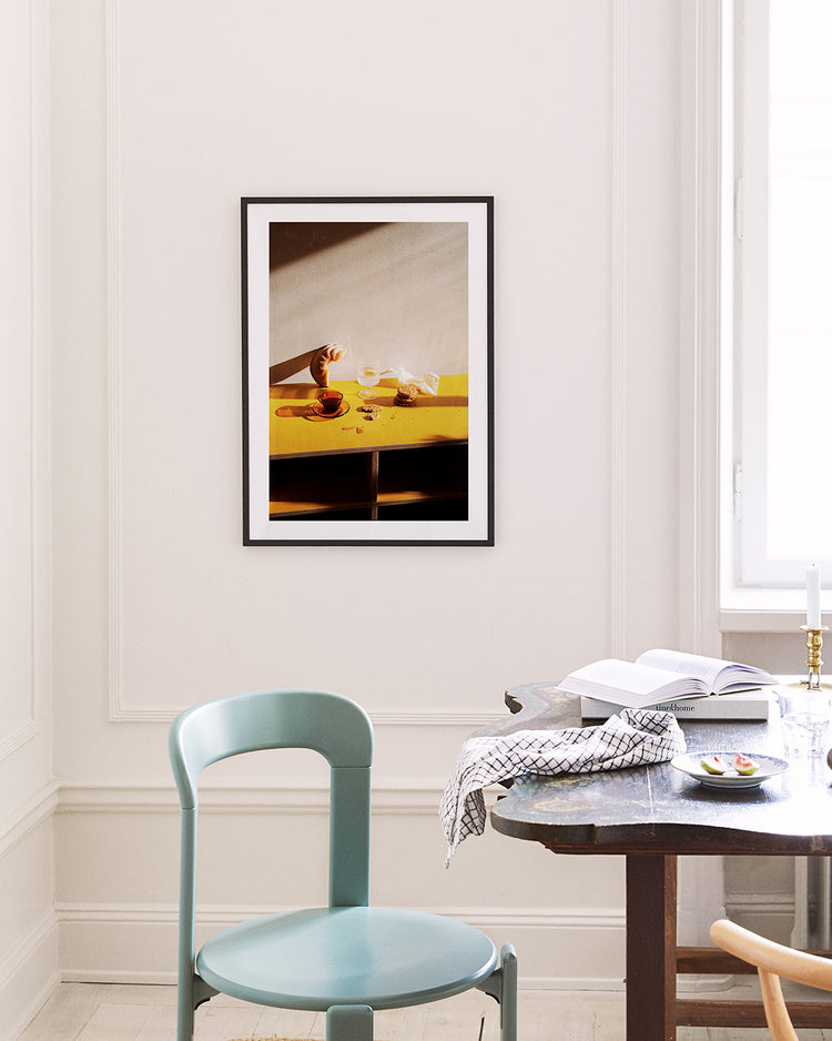 A minimalist room features a Would You Like Some Coffee? poster on a white wall, showing a hand pouring liquid into a cup on a yellow table. Nearby are a blue chair and dark wooden table with an open book and patterned cloth, all bathed in natural light.