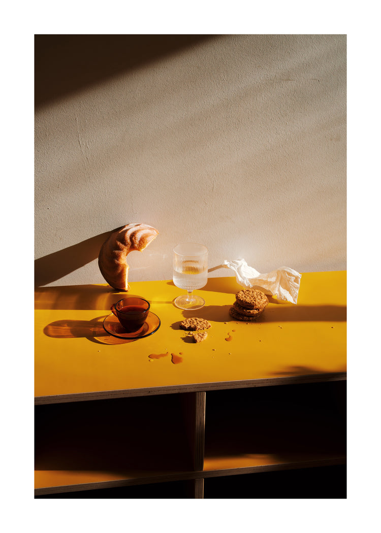 On the yellow table, a croissant, a cup of tea, cookies, and a glass of water sit alongside a folded napkin. Crumbs are scattered about. Soft lighting creates welcoming shadows reminiscent of the ambiance captured in the Would You Like Some Coffee? poster.