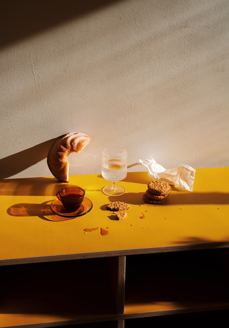 The Would You Like Some Coffee? Poster illustrates a still life with a croissant against a wall, glass of water, cup of tea, cookie crumbs, stacked cookies, and crumpled napkin on a bright yellow surface.
