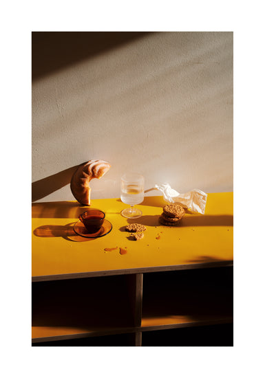 The Would You Like Some Coffee? poster depicts a croissant leaning against a wall on a yellow table beside a glass of water, a tea cup, and three cookies. Sunlight creates inviting shadows, while a crumpled napkin lies near the cookies.