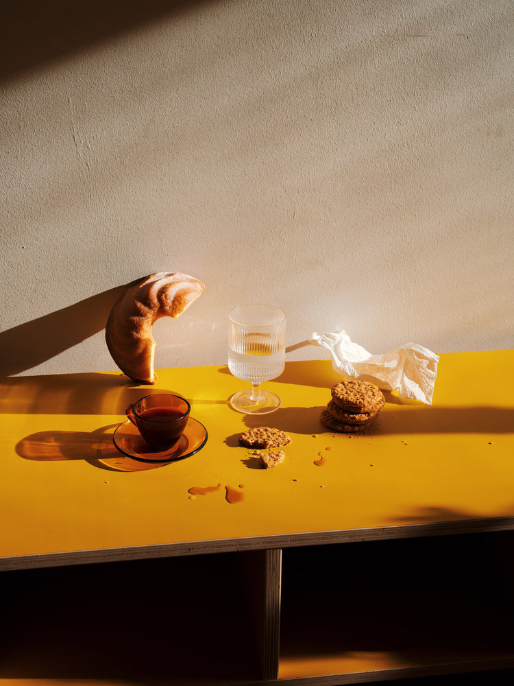 The Would You Like Some Coffee? poster depicts a fresh croissant, glass of water, cup of tea, scattered cookies on a yellow table, and a crumpled napkin nearby. Sunlight casts shadows that add warmth to the scene.