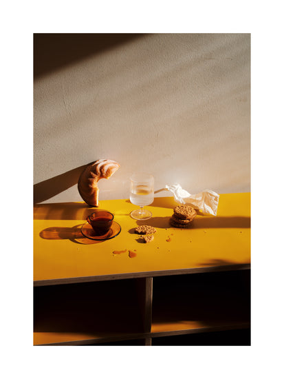 The Would You Like Some Coffee? poster depicts a sunlit scene with a croissant on a yellow table, beside a glass, dark liquid in a cup, a crumpled napkin, and cookies. Shadows enrich the wall and table, crafting warmth and an inviting aura.