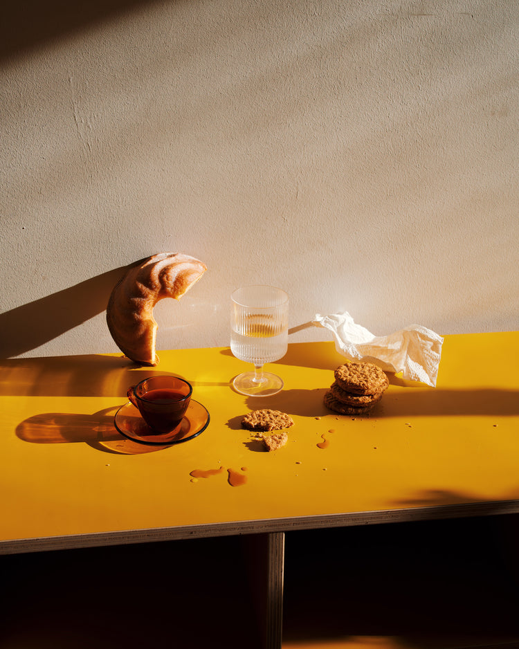 The Would You Like Some Coffee? Poster features a croissant resting against a wall on a yellow tabletop alongside a glass of water, teacup, and cookie crumbs. Bathed in bright sunlight, the scene creates an inviting and warm ambiance with long shadows.
