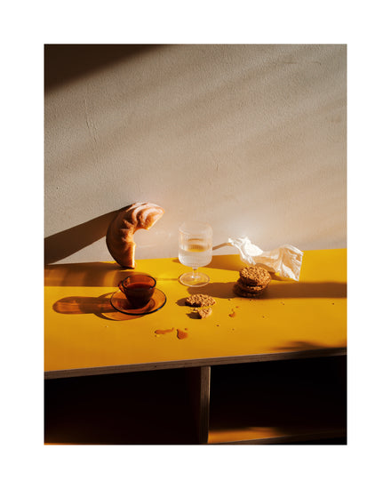 The Would You Like Some Coffee? poster shows a warmly lit yellow table with dramatic shadows, featuring a croissant, crumpled napkin, glass of water, and a small tea cup with tea. Crumbs are scattered across the surface.