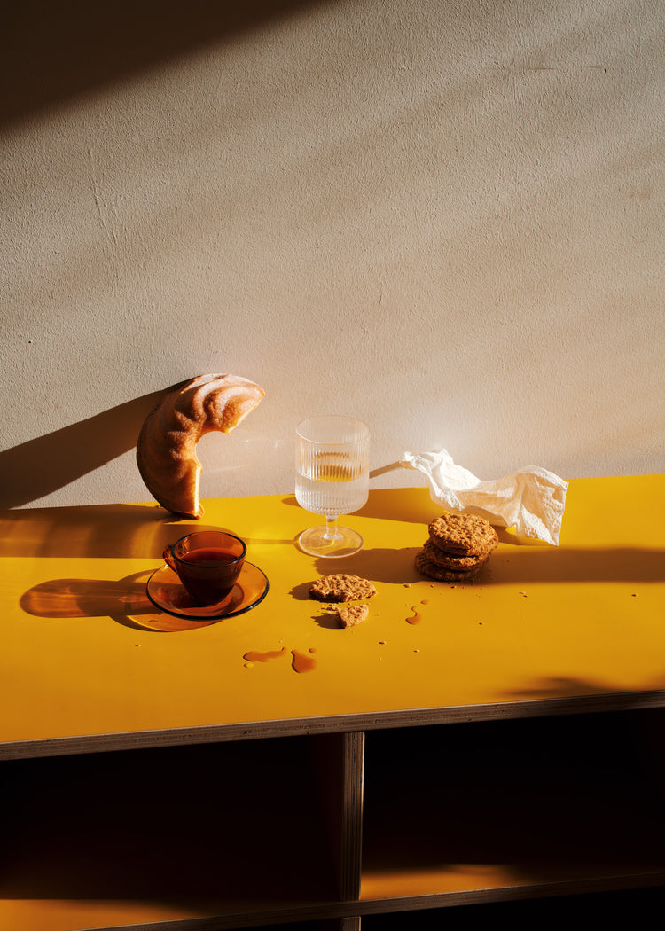 Under the sunlight, creating a warm scene, the yellow table displays a croissant, cookies, tea and water glasses, and a crumpled napkin. This inviting arrangement is featured in the Would You Like Some Coffee? poster against a plain wall.