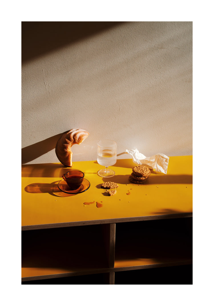 The Would You Like Some Coffee? Poster depicts a golden table adorned with a croissant, a glass of liquid, a teacup, a crumpled napkin, and cookie crumbs. Sunlight casts warm shadows, creating an inviting scene.